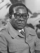 Robert Mugabe, wearing a dark suit and glasses, looks to the viewer's right