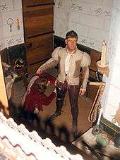 A wax figure of a man stands, preparing for battle. The figure sports a bowl cut and wears a khaki doublet and dark brown breeches. At its legs is the wax figure of a kneeling squire.