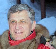 A man with grey hair, wearing a red and brown coat, with snow in the background.