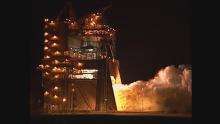A video showing RS-25 testing. The video opens with a night view of a large scaffold structure (the test stand), lit with internal lights. The view then switches to show the nozzle of a rocket engine, mounted within the structure, lighting and beginning to fire. The view then cuts back to the view of the scaffold, from which large amounts of steam are now billowing out of, towards the right of the frame. Wide and close-up views of this plume follow, before the view switches back to the engine nozzle, which shuts down.