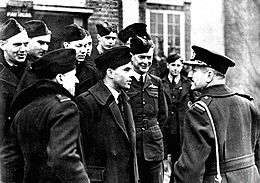 Half-length outdoor portrait of moustacioed man in military great coat with peaked cap, talking to a group of ten or men in military uniforms with forage caps