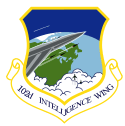 A standard shield, surrounded on the edge by a diminished yellow border. The upper part of the shield is blue, with one gray aircraft with a white trail pointing out from the left side. The plane is flying over the northeastern portion of the western hemisphere, which is colored in green and light blue. On the front of the gray aircraft with a white outline is a cockpit, with the jet firing three rockets. The three rockets trail white smoke and are aimed the same direction as the aircraft. Under the nose of the plane are two black aircraft coming out of clouds and rising up towards the jet. Attached to the shield is a white scroll edged with the same color border as the shield, surrounding the inscribed "102D INTELLIGENCE WING" in blue