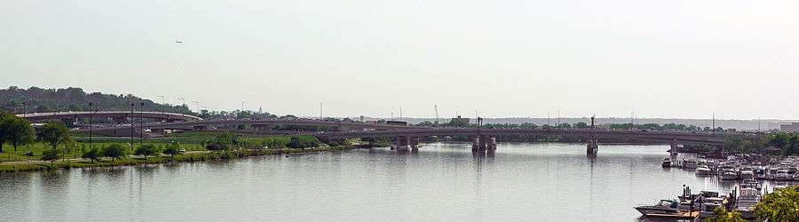 Photograph of a bridge gently curving over a wide river, with a marina to the right and flyovers to the left.