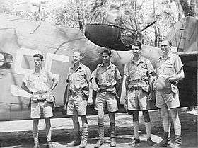Five men in tropical military uniforms stand beside rear fuselage and turret of Lockheed Hudson bomber