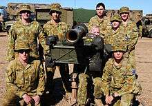 Photograph of eight people wearing camouflage uniforms posing with a rocket launcher. Several camouflaged trucks are visible in the background.