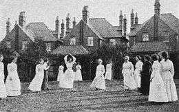 A group of 15 women in long-sleeved shirts and ankle-length skirts on a grass netball court.