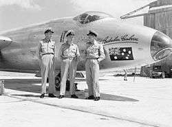 Black and white photograph of three men wearing military uniforms posing while standing in front of the nose of a military jet aircraft