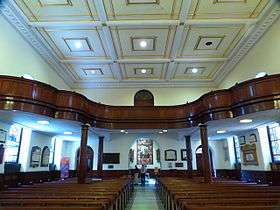 The interior looking towards an arched exit. There are rows of pews and a wide centre aisle. The ceiling is coffered into decorative square shapes. There is a large gallery of dark wood with a curving balustrade across the back of the church.
