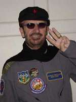 A colour photo of Starr, who is holding two fingers up in the style of a "peace sign". He is a wearing a dark beret, sunglasses and a grey windbreaker with several patches on the front.