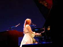 A woman with red hair in a white dress, playing a piano and singing into a microphone.