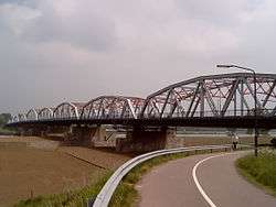 A steel bridge with red inner supports spans a dry canal and a river; the bridge is fronted by a road seen in the front of the image.