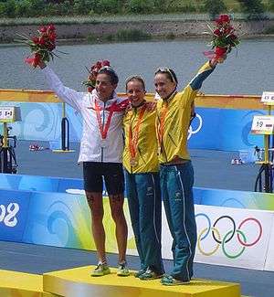 Three women wearing medals around their necks stand together on the highest step of a podium. The woman on the left wears dark blue shorts, a white jacket and raises a flower bouquet with her right arm. The other women wears the same turquoise pants and yellow jackets; the one on the right also holds high a flower bouquet with her left arm. Far behind them is a large water source and vegetation.