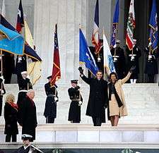 The Obamas wave as they walk past military flagbearers in the background while a couple stands in the foreground.