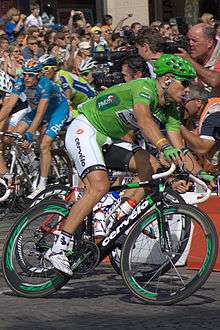 The same cyclist in the green jersey bearing the Cervélo logo. He again wears a green helmet and sunglasses with green frames, and his bicycle has special green accents in it. Behind him are riders from other teams, as well as many spectators watching from the roadside.