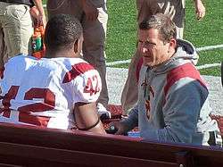 Candid photograph of Berry wearing a hooded sweatshirt with a USC logo and kneeling on a football sideline and speaking with a player seated on a bench