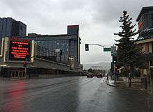 View of a rain-soaked roadway passing between tall hotel-casino buildings on either side and a traffic signal in the foreground, with signs indicating the Nevada state line at Lake Tahoe