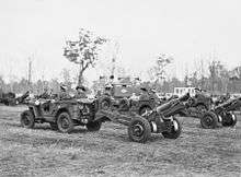 Small motor vehicles towing artillery guns across a grassed area. Trees are visible in the background.