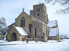  Stone building with gravestones in foreground and to the left; the church tower is surmounted by an ornamental weathercock, and has a clock showing 10.35. The roofs and surrounding areas are covered in snow.
