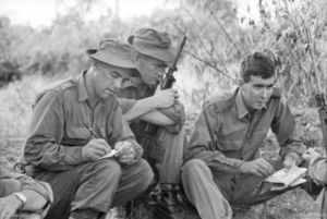 A soldier holding a map is talking while two other soldiers sitting nearby look on while taking notes.