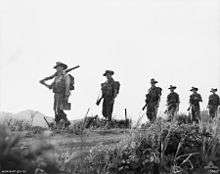 Six evenly spaced Caucasian soldiers wearing slouch hats with their rifles slung march along a dirt road in a straight line from right to left, carrying packs. The camera is at ankle height amidst the grass which is in the foreground, while the men are silhouetted against the skyline.