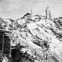 The snow covered ridgeline of a steep hill running away from the camera and sloping downwards from left to right. In the bottom left foreground two soldiers with machine-guns stand in a trench in front of a defensive position constructed of sandbags and logs, facing downhill. In the background several trees, stripped of foliage, stand on crest.