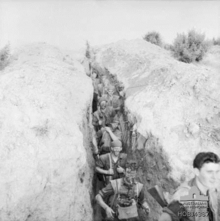 A deep trench line runs vertically from the top to the bottom of the photography, while on either side is a bare earth bund. Nine Caucasian soldiers carrying weapons, ammunition, and other equipment, many with their faces blacked and wearing camouflage on their heads, move in a straight line down the trench towards the bottom of the photo, below the camera. At the bottom right of the photo, a soldier stands level with the camera facing the right at the head of the patrol.