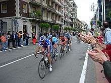 Various cyclists, clad in jerseys of different colors, speed down an open road. Spectators watch from the roadside, and cheer them on