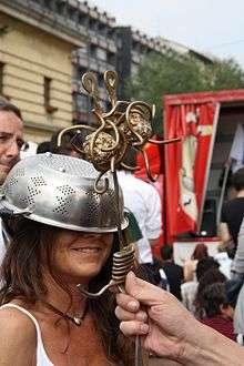 smiling woman wearing a colander on her head being "blessed" by a brass Flying Spaghetti Monster in the style of a Roman Catholic scepter.