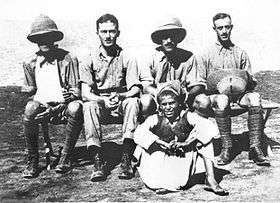 Four men in khaki military uniforms, two wearing pith helmets (one of whom has his arm in a sling), seated in front of a boy