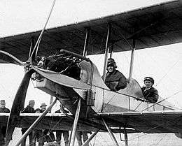Two men in flying gear seated in tandem open cockpits of a biplane with a four-bladed propeller