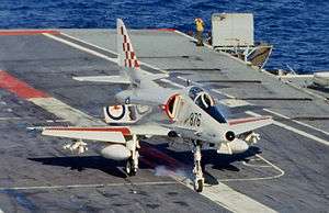 A jet aircraft with red and white markings and the word "Navy" on its tail lands on the grey deck. Smoke rises from the front tire. Water can be seen in the background.