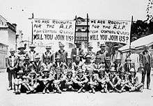 Black and white group portrait of a group of men wearing suits seated and standing in front of two banners. Both banners read "We are the recruits for the A.I.F. from Central Q'Land [Queensland] will you join us?" in block letters.