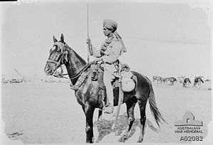 In a black and white photograph, a man wearing a turban and light-coloured tunic sits astride a dark-coloured horse facing left and holds a bayonet in his right hand. He is depicted against a desert landscape.
