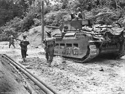 An armoured vehicle moves along a sandy road surrounded by jungle. Infantrymen advance alongside