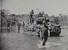 A tank moves across a shallow creek with armed soldiers advancing beside it