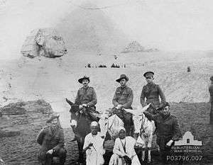 Men wearing military uniforms sitting atop horses with others in local dress in front of pyramids