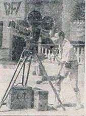 A man with short hair and a white shirt is controlling a large camera. He is facing left.
