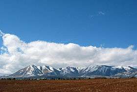 Snow-covered mountains protruding from a plain with tilled soil in the foreground.