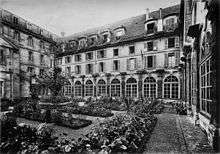 A black-and-white photograph taken from the vantage point of a person standing in the rectangular courtyard at Abbaye-aux-Bois. The grounds have rectangular plots of small plants, surrounded by grass cut very short. A brick path lines the walls of the four-storey building surrounding the courtyard.