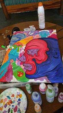 An acrylic painting of a red and blue face in profile lies on top of a tray on a table. Below the painting, on the table, are a palette and various sizes and colors of paint.