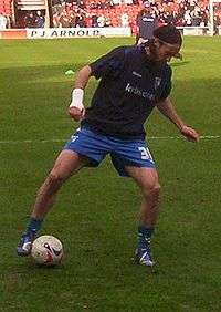 A man wearing a black t-shirt and blue shorts standing on a grass pitch, with a spherical football ball next to one of his feet.