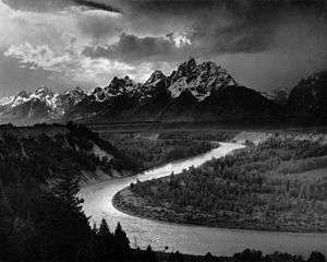 A dramatically-lit black-and-white photograph depicts a large river, which snakes from the bottom right to the center left of the picture. Dark evergreen trees cover the steep left bank of the river, and lighter deciduous trees cover the right. In the top half of the frame, there is a tall mountain range, dark but clearly covered in snow. The sky is overcast in parts, but only partly cloudy in others, and the sun shines through to illuminate the scene and reflect off the river in these places.