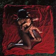 An aerial view of a nude black woman sitting on a red blanket with a white baby held in her arms.