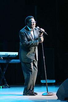 A man standing behind a microphone stand on a stage, wearing a suit, bow-tie and glasses