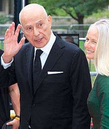 A Caucasian male wearing a black suit and tie with a white collar shirt is seen waving his right hand. He is standing next to a blonde female wearing a green dress.