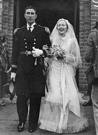 A young couple in wedding attire walking outside of a building. The man is on the left, and is dressed in ceremonial naval attire. He is holding a hat in his right hand down by his side. The bride is in a white gown with a veil. She is smiling and carrying a bouquet of flowers