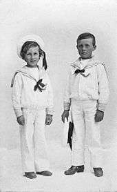 A black and white portrait of two young boys. They are standing alongside one another and are dressed in white sailor uniforms. The shorter boy on the left is smiling and wearing a cap, while the boy on the right has a blank expression and is holding a cap in his left hand.