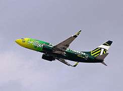 Left side view of a mostly-green aircraft with Portland Timbers colors taking off in clear skies