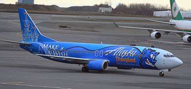 An aircraft painted dark blue with a genie on the fuselage and a castle on the tail, taxiing on the tarmac with another aircraft and some trees in the background.