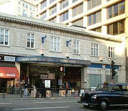 A light grey building with "M.R. ALDGATE STATION M.R." written in stone on the front face and a black car driving in the foreground